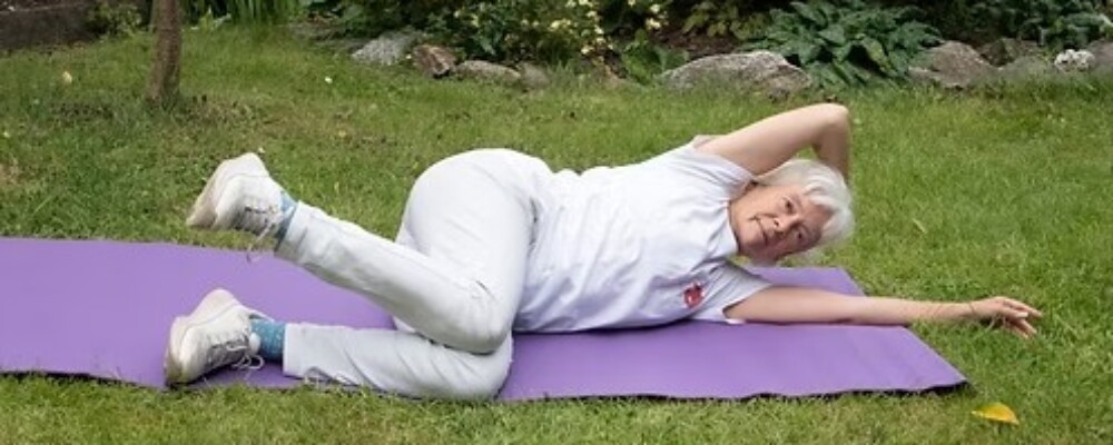 Photograph of ninja granny laying on a yoga mat on some grass. She is doing a somatic exercise laying on her side.