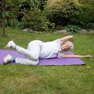 Photograph of ninja granny laying on a yoga mat on some grass. She is doing a somatic exercise laying on her side. 
